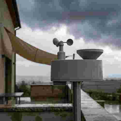Wetterfühler an Metallmast bei Regen und Wind neben Schattensegel, mit Gewitterwolken im Hintergrund